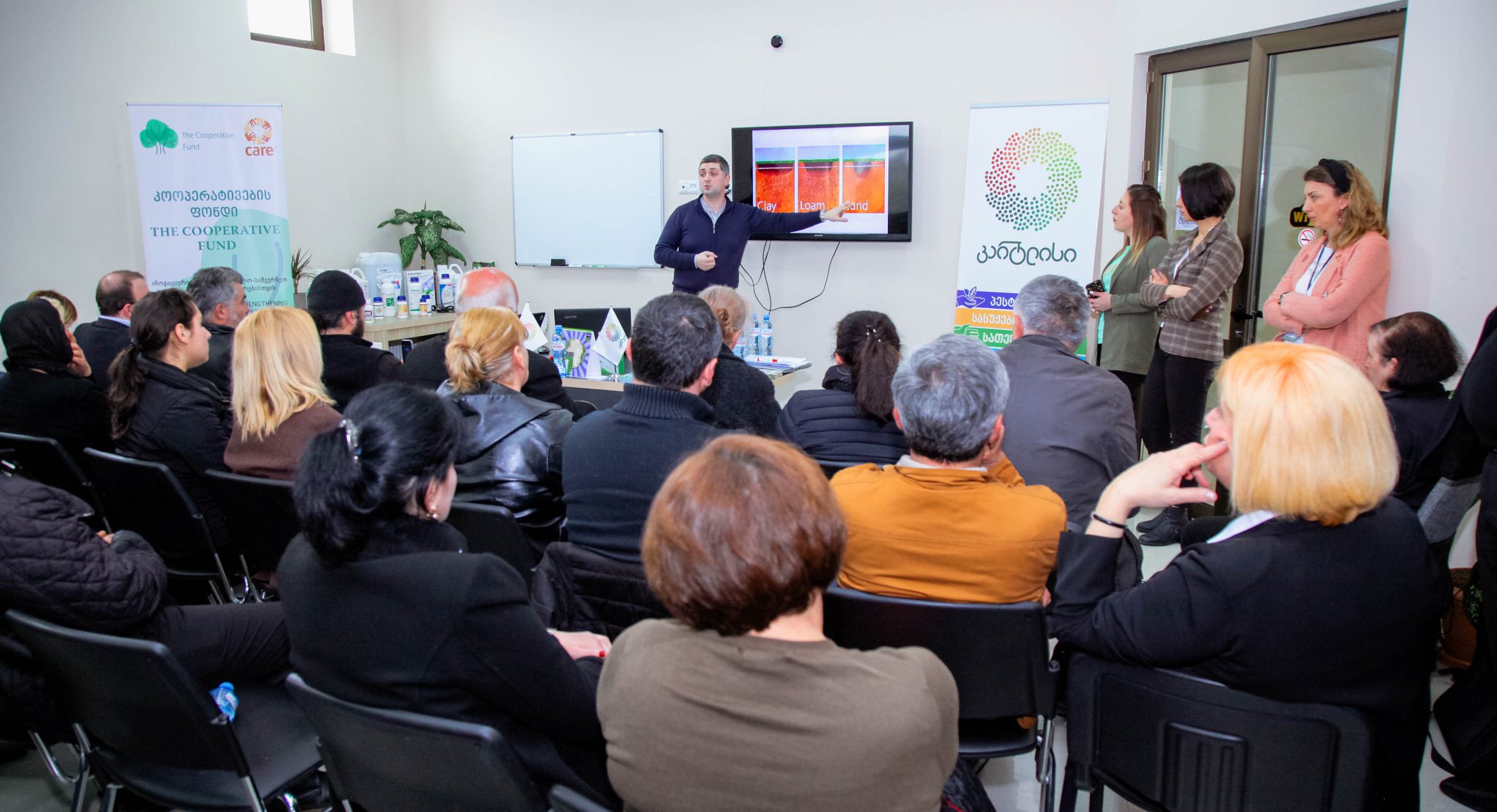 A group of people sit in a meeting with their attention focused on a presenter standing in front.