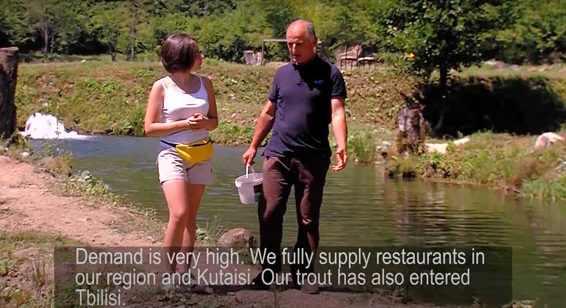 A man and woman walk down a narrow path along a pond. He is talking and holding a white bucket while she listens.